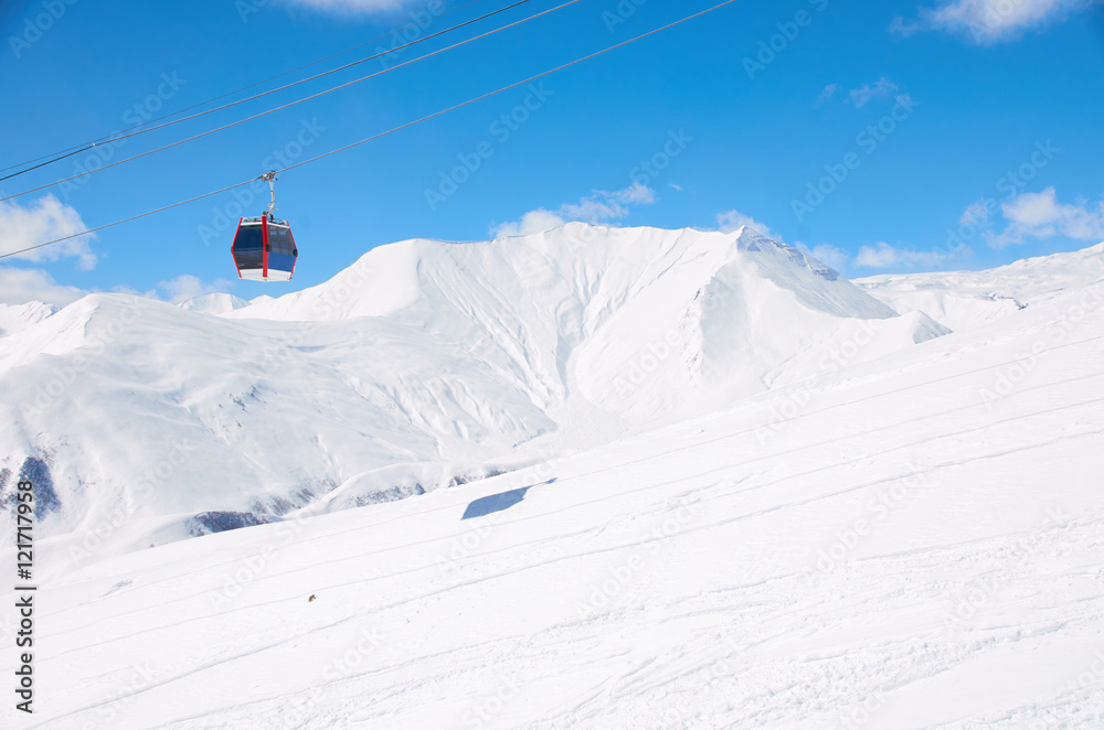 Cable lift for skiers or snowboarders in brigth day on ski resort with blue sky and white snow mountain picks background. Copy space for a text. Red car or cabinet for athletes.