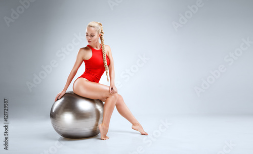 High fashion portrait of a slim and beautiful fitness model with blonde hair posing in a red bodysuit on a grey background