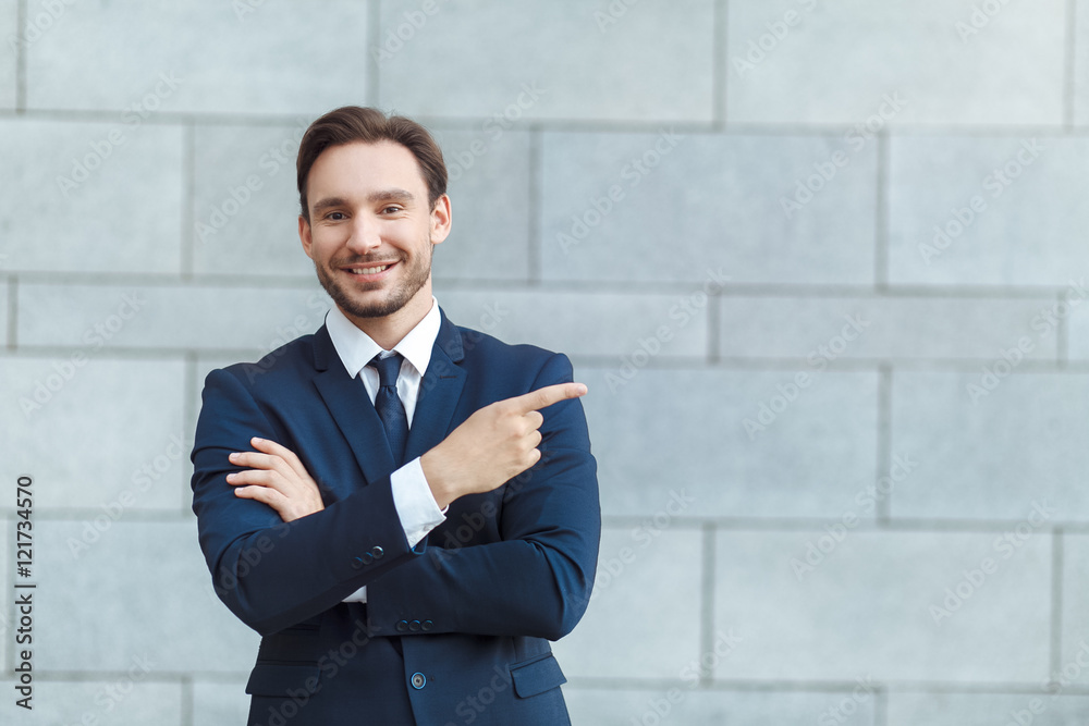 Look over there! Young handsome businessman in formalwear pointing away and smiling while standing in front of the grey wall