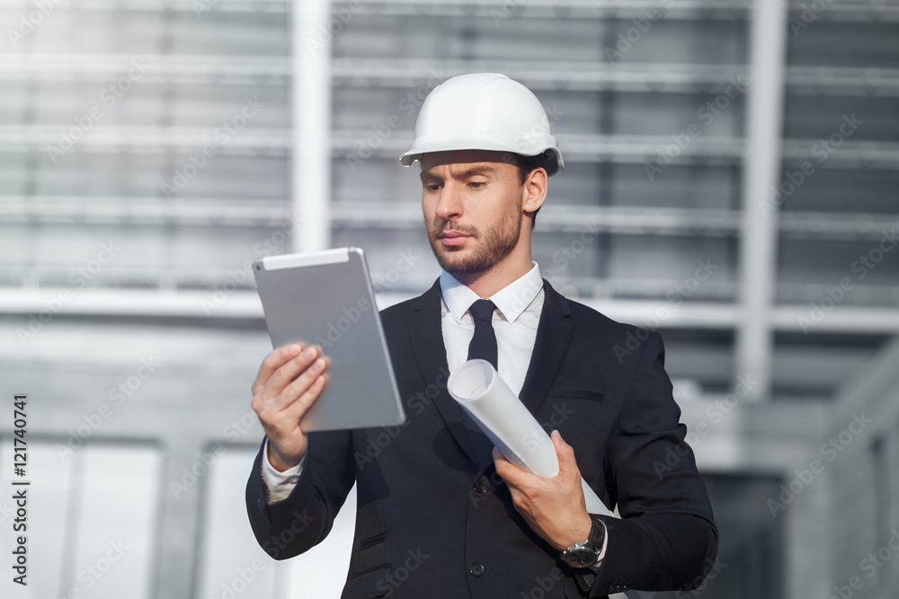 Confident architect in hardhat holding blueprint and working on laptop while standing on background of building structure
Confident architect in hardhat holding blueprint and working on laptop while standing on background of building structure
