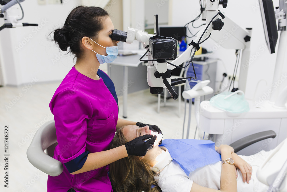 Young female dentist treating root canals using microscope at the dental clinic. Young woman patient lying on dentist chair with open mouth. Dentist wearing mask and gloves Young female dentist treating root canals using microscope at the dental clinic. Young woman patient lying on dentist chair with open mouth. Dentist wearing mask and gloves