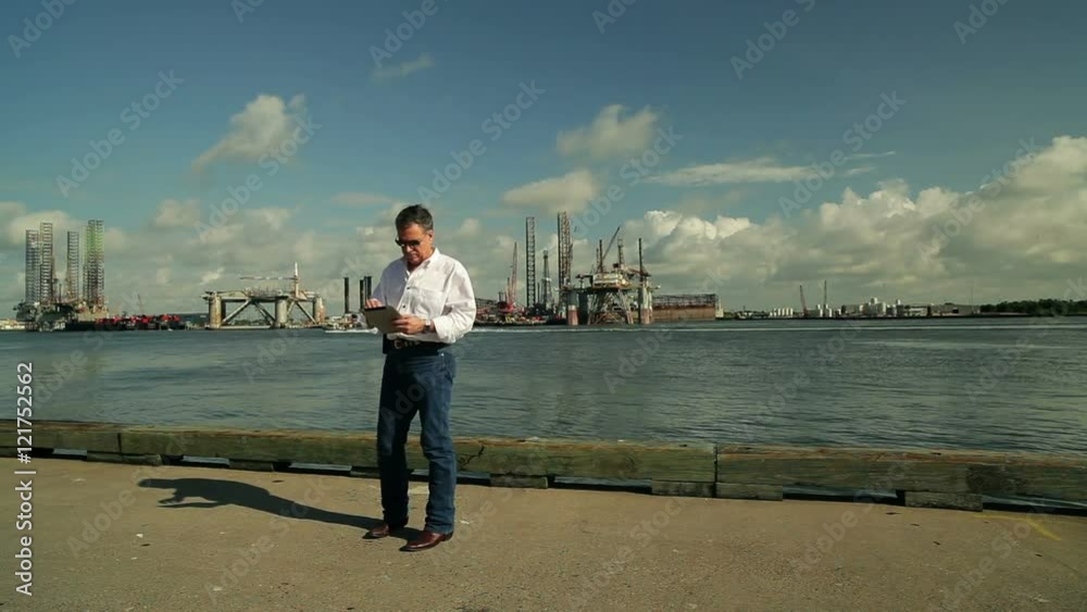 Drilling platforms and other equipment from the oil and gas industry are the background for a man on a dock using an electronic tablet. Drilling platforms and other equipment from the oil and gas industry are the background for a man on a dock using an electronic tablet.