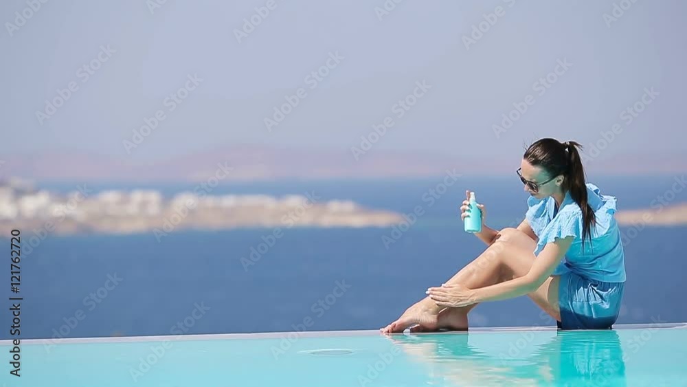 Slim woman applying sunscreen on her legs, sitting on the edge of pool background old town Mykonos in Europe Slim woman applying sunscreen on her legs, sitting on the edge of pool background old town Mykonos in Europe