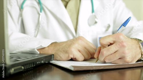 The camera dollies past a laptop computer to a left handed doctor writing on a chart or patient record.