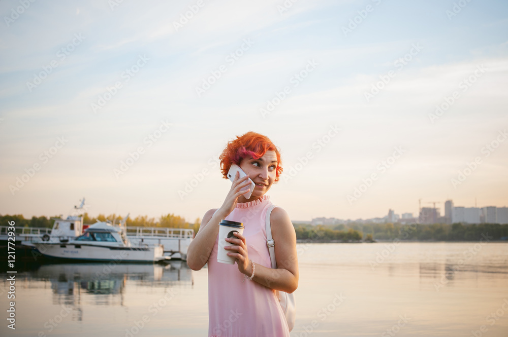 girl in pale pink dress with red hair and backpack walking along river bank, talking on the phone and drinking coffee from a cardboard cup, against backdrop of boats moored on a warm summer day