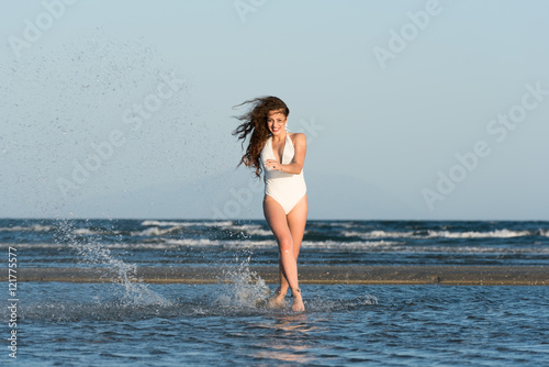 Woman with long curly hair and excellent body wear white monokini, running in the water. Sea and sky as background