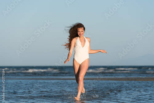 Woman with long curly hair and excellent body wear white monokini, running in the water. Sea and sky as background