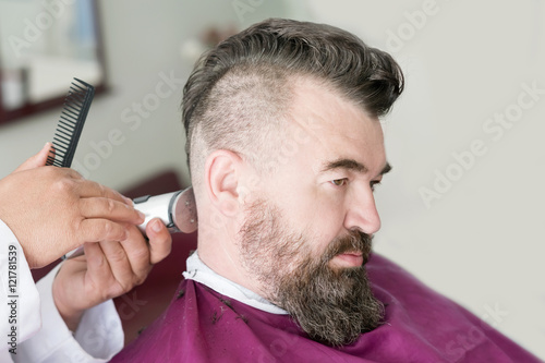 Male barber makes a mohawk hairstyle using clipper of a adult man with beard photo