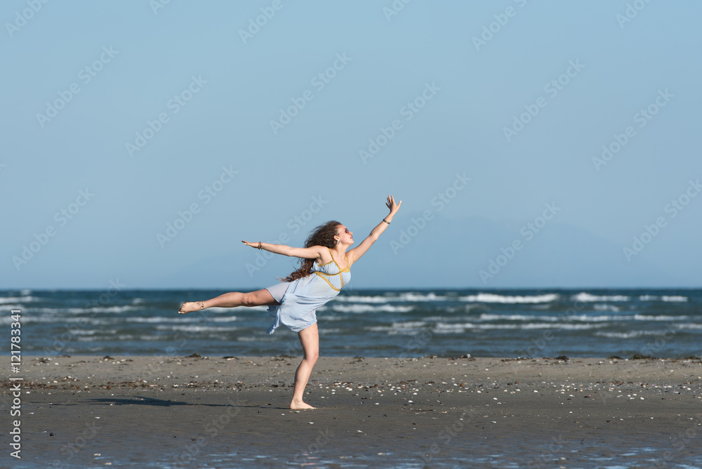 Woman with long curly hair wear dance dress and making moves, standing on the beach. Wind wave the skirt. Sea and sky as background