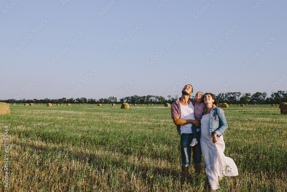 father in a plaid shirt and mother in a denim jacket and white dress hug her little daughter outdoors in field and looking up