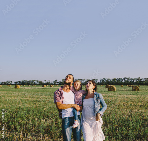 father in a plaid shirt and mother in a denim jacket and white dress hug her little daughter outdoors in field and looking up