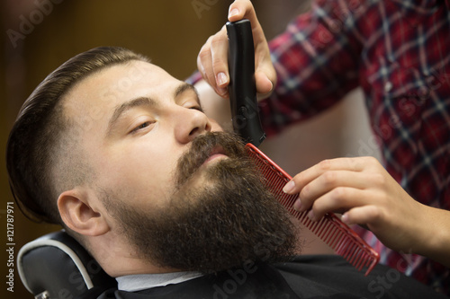 Close-up portrait of handsome young bearded caucasian man getting trendy beard haircut in modern barber shop. Hairstylist girl working, serving client, doing beard grooming using shaver and comb