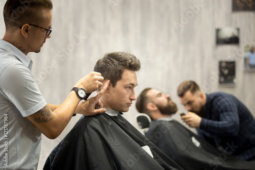 Interior shot of working process in barbershop. Side view of handsome young men getting trendy haircuts in modern barbershop. Cool male hairstylists serving clients