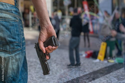 Gun control concept. Armed man - attacker holds pistol in hand in public place. Many people on street. photo