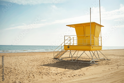 Footprints and wheeltracks on the sand beyond an empty yellow lifeguard cabin on a deserted beach © BublikHaus