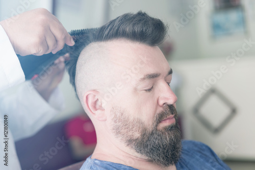 Male barber makes hair styling using a hair dryer of a adult bearded man with a mohawk