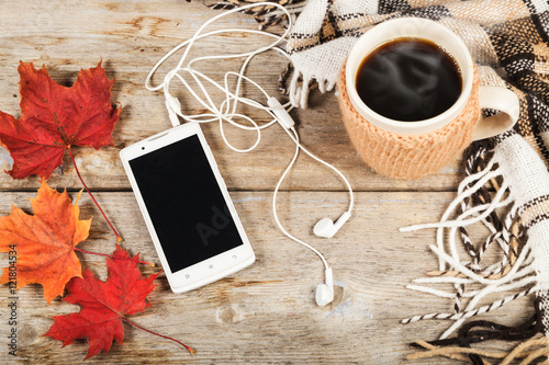 Hot coffee in a large cup, white mobile phone with headphones, three maple leaf, checkered plaid on a wooden table surface