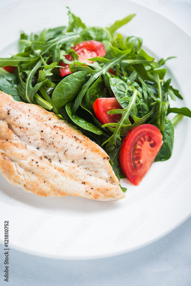 Roasted chicken breast and fresh salad with tomato and greens (spinach, arugula) close up on white textured background. Healthy food.