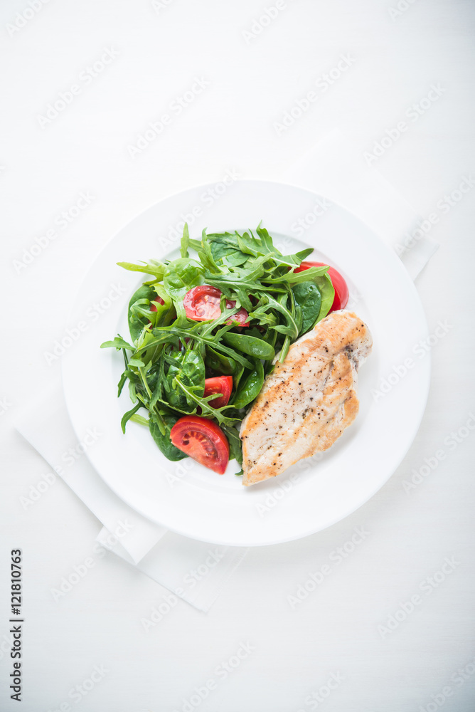 Chicken breast and fresh salad with tomato and greens (spinach, arugula) top view on white wooden background. Healthy food.