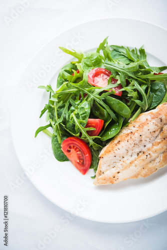 Roasted chicken breast and fresh salad with tomato and greens (spinach, arugula) top view on white textured background. Healthy food.