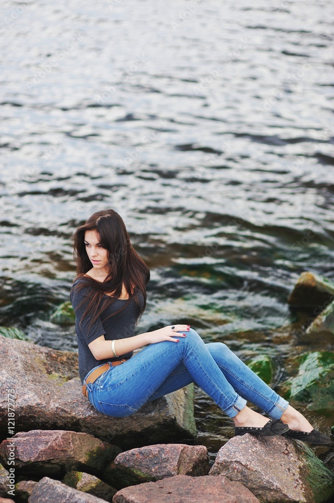 Young beautiful brunette girl with long hair sitting on the rocks by the sea  a background of dark green waves.