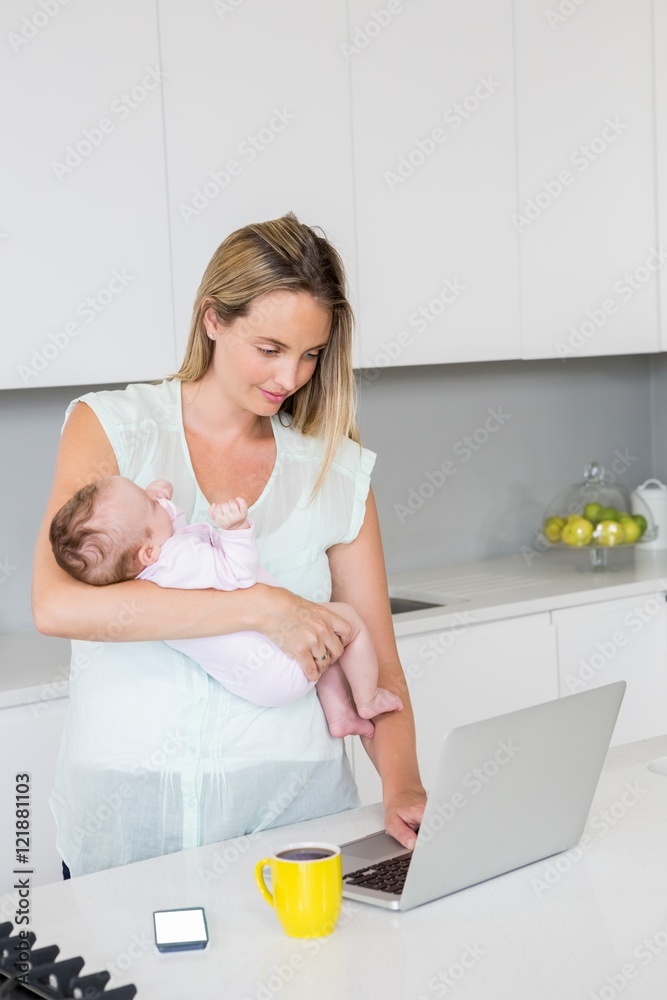 Mother using laptop while carrying baby in kitchen Mother using laptop while carrying baby in kitchen