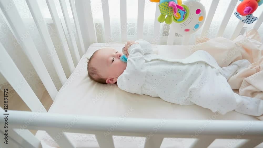 Dolly shot of cute 3 months old baby boy lying in white wooden crib with toy carousel