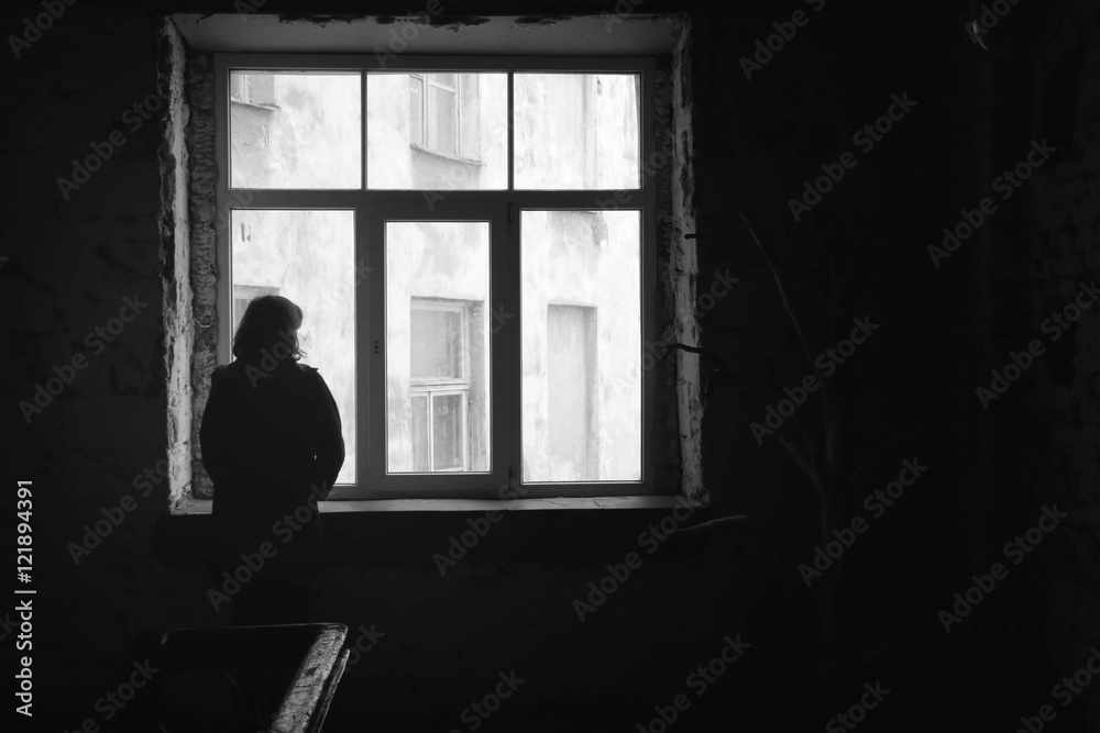 Concept of loneliness in the city, waiting and decision making. Woman looking out of the window in dark deserted house, Selective focus. Special light.