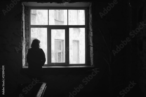 Concept of loneliness in the city, waiting and decision making. Woman looking out of the window in dark deserted house, Selective focus. Special light.