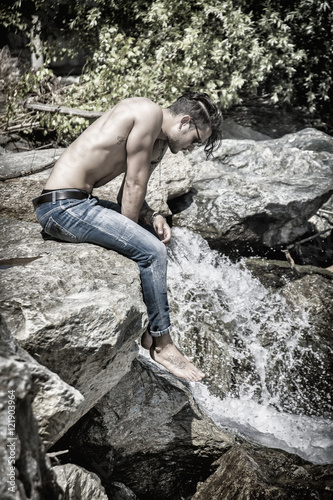 Athletic shirtless young man outdoor at river or water stream, sitting on big rock, looking away, with stones in background