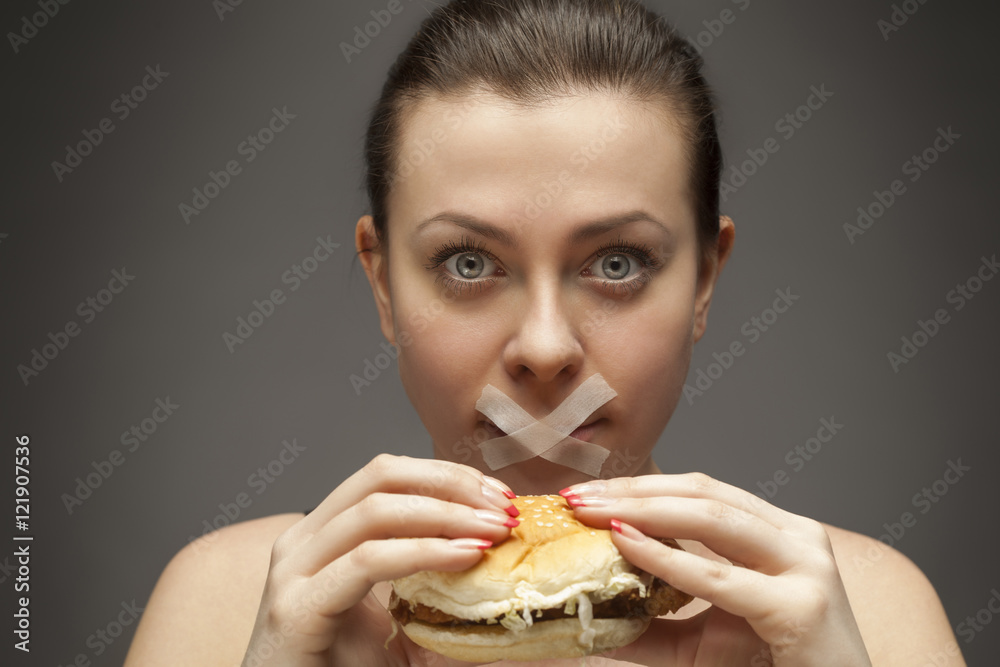 Diet concept: woman holding a burger with mouth sealed Diet concept: woman holding a burger with mouth sealed