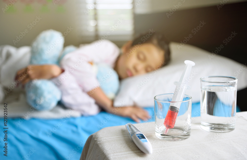 Generic liquid medicine syringe in glass on table in front of out of focus sleeping child for health and illness concept, young Asian girl hugs the blue teddy bear. Generic liquid medicine syringe in glass on table in front of out of focus sleeping child for health and illness concept, young Asian girl hugs the blue teddy bear.
