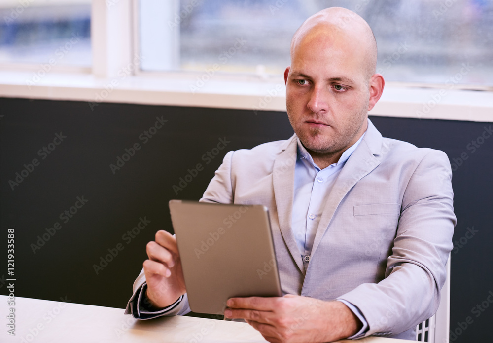 Business man holding and using a high tech tablet while seated at a conference table early in the morning with a window behind him and light coming in through the glass. Business man holding and using a high tech tablet while seated at a conference table early in the morning with a window behind him and light coming in through the glass.