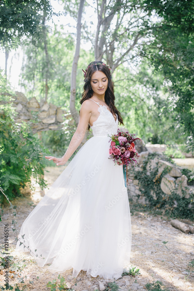 portrait of a beautiful happy brunette bride in wedding white dress holding hands in pink and purple bouquet of flowers outdoors on the background of green leaves