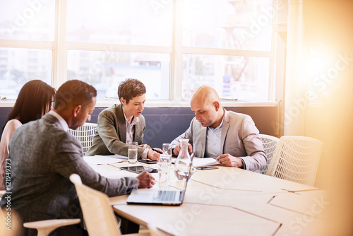 Four business professionals conducting a meeting in a bright modern conference room with a large windown and an abundance of natural light, while making use of technology to optimise efficiency.