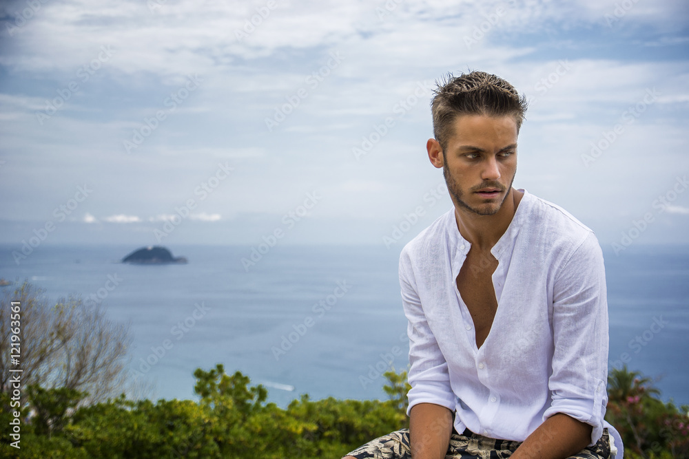 Handsome Young Man in Trendy Attire, n a Sunny Summer Day with Italian Sea Coast in the Distance, Wearing a White Shirt