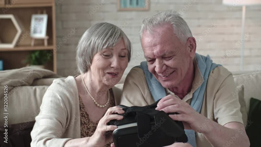 Laughing elderly couple sitting on sofa and looking with curiosity into virtual reality headset, then trying them on and checking if they can see in them Laughing elderly couple sitting on sofa and looking with curiosity into virtual reality headset, then trying them on and checking if they can see in them