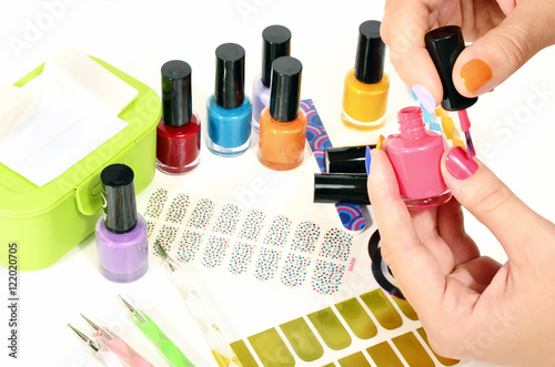 Woman hands doing rainbow manicure.Table full of manicure ustensils,coloured nail polish,nail decals stickers,striping tape,nail file,dotting pens,vinyl sheets.Nail art accesories isolated on white. 