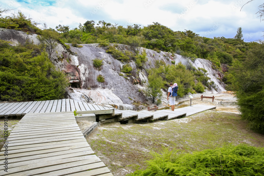 Fototapeta premium Young active father with little daughter in a back carrier standing on wooden walkboard and observing Thermal area Orakei Korako in New Zealand