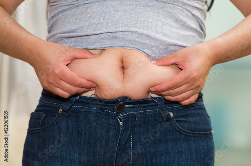 Closeup womans stomach wearing jeans, grabbing onto excessive fat using fingers, belly button revealed, weightloss concept