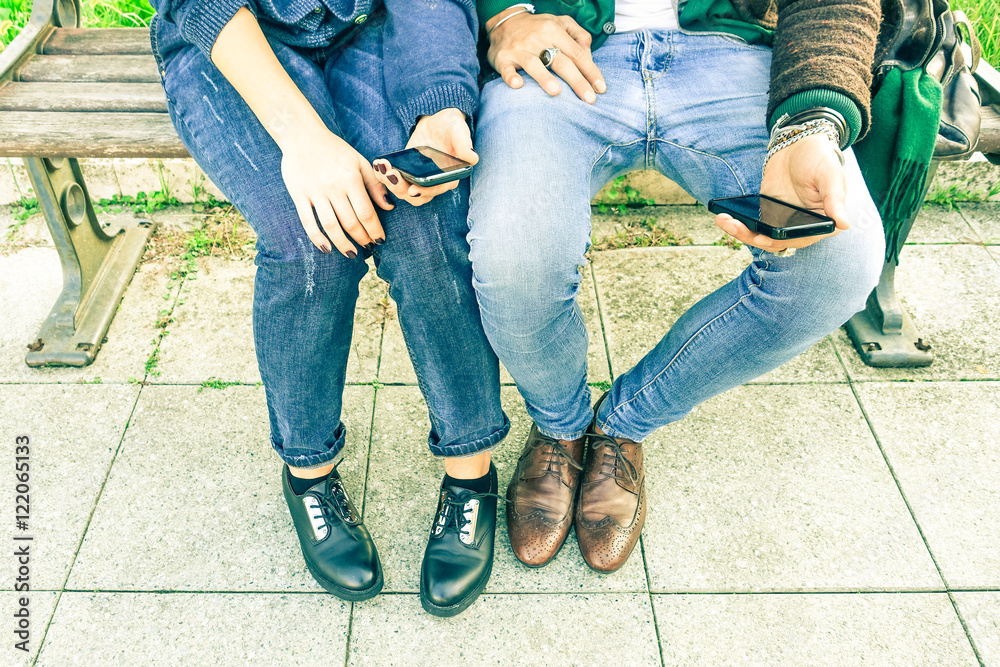 Couple of hipsters holding mobile phone sitting on bench - Two trendy guys using cell at city park - Modern concept of romance Couple of hipsters holding mobile phone sitting on bench - Two trendy guys using cell at city park - Modern concept of romance