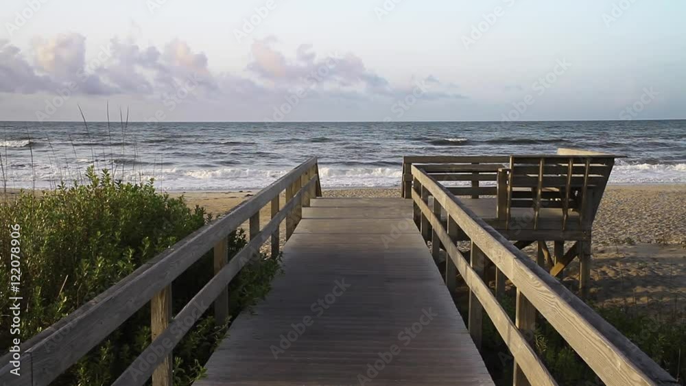 Loop features ocean waves breaking on the shore under a morning sky with puffy clouds as viewed down a boardwalk leading to the beach. Shot on North Carolina's Ocracoke Island.