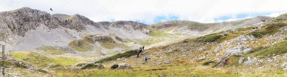 panoramic photo of a valley in Abruzzo, Italy. At the center, some hikers climb the valley, on the right and left some deer observe an eagle flies over the mountains.