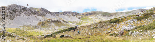 panoramic photo of a valley in Abruzzo, Italy. At the center, some hikers climb the valley, on the right and left some deer observe an eagle flies over the mountains.