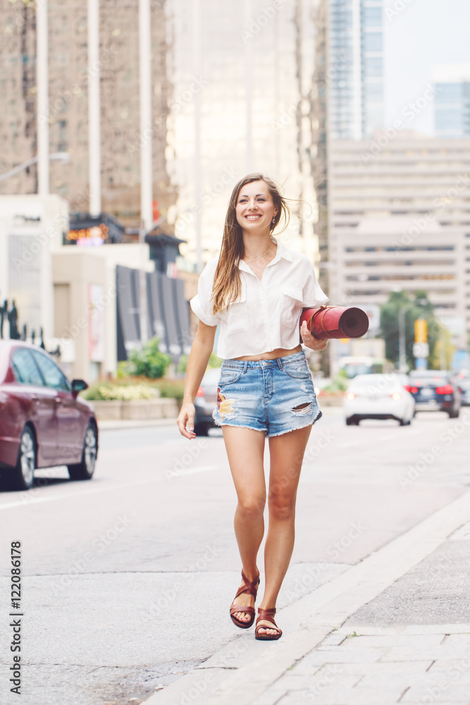 Portrait of smiling happy slim fit sporty young white Caucasian hippie hipster woman with long blond hair going with yoga mat on busy city street among cars, healthy lifestyle concept.