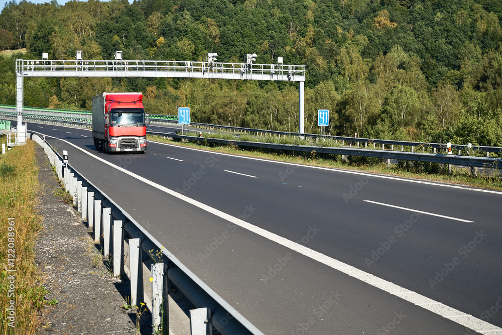 Electronic toll collection with a passing red truck on a highway on the forest background.