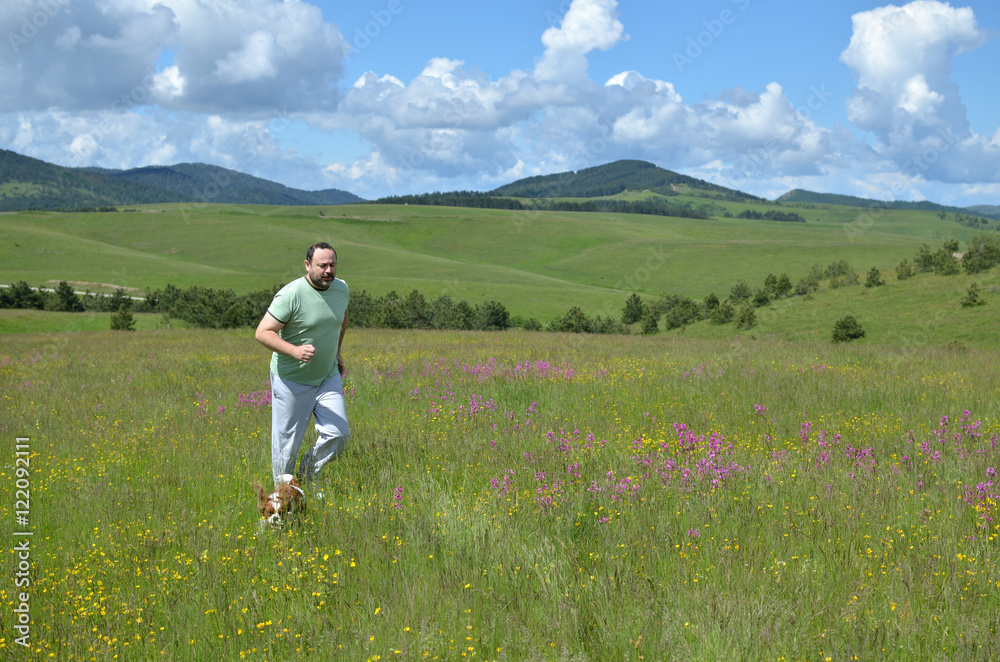 Man and his dog running in a field with wildflowers on a cloudy day in springtime and with hills in background
