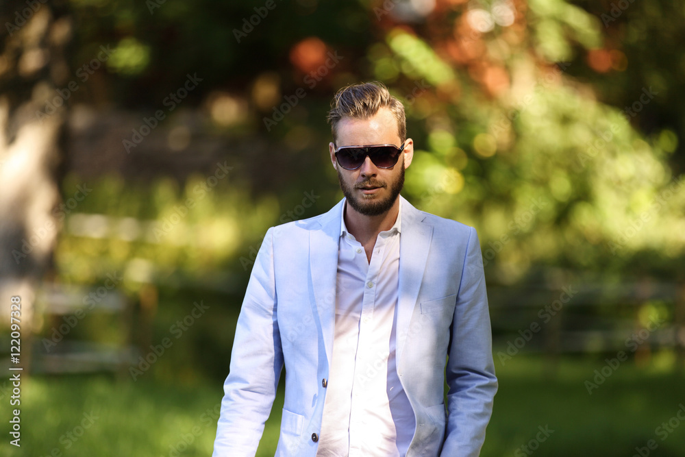 An attractive man wearing a light blue blazer, white shirt and sunglasses,  walking along a path outside on a sunny summer day. Stock Photo