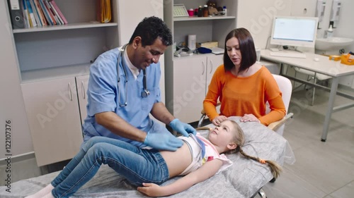 Latin-American doctor performing abdominal examination on little girl with her smiling mother sitting beside