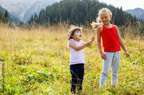Little Asian girl walking with caucasian girl on meadow with mountains in the background